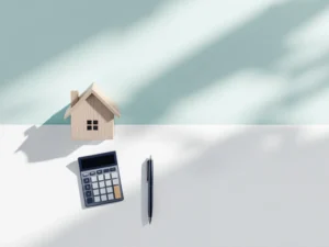 Houten huismodel met rekenmachine, pen en aflopende stapels munten op witte ondergrond, symboliseert hypotheekreductie