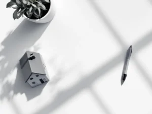 Bovenaanzicht van wit bureau met architectuurmodel van huis, rekenmachine, pen en weegschaal icoon in grijstinten